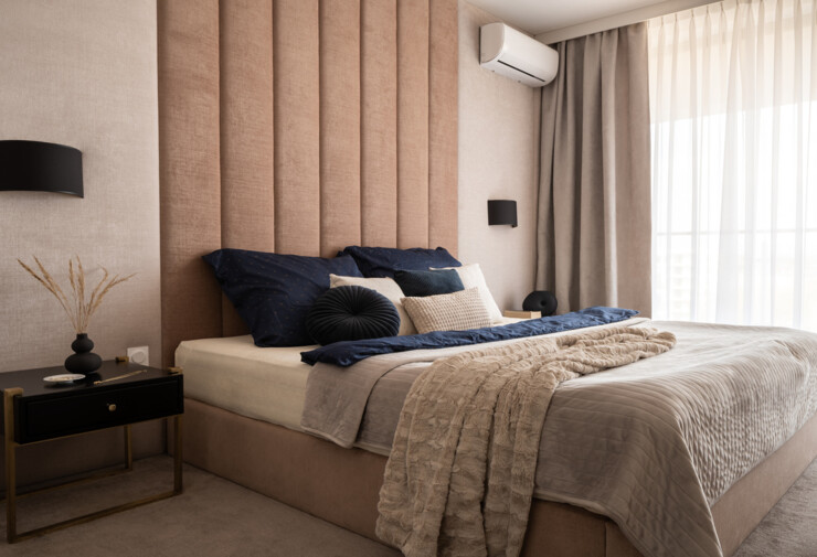 Climatisation discrète murale installée en hauteur dans une chambre moderne aux tons beiges, intégrée près du plafond au-dessus des rideaux, avec lit double élégant et décoration minimaliste.