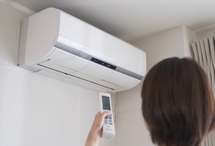 Climatiseur mural réversible blanc installé en hauteur dans un salon lumineux, avec une personne utilisant une télécommande pour ajuster la climatisation et le confort thermique intérieur.